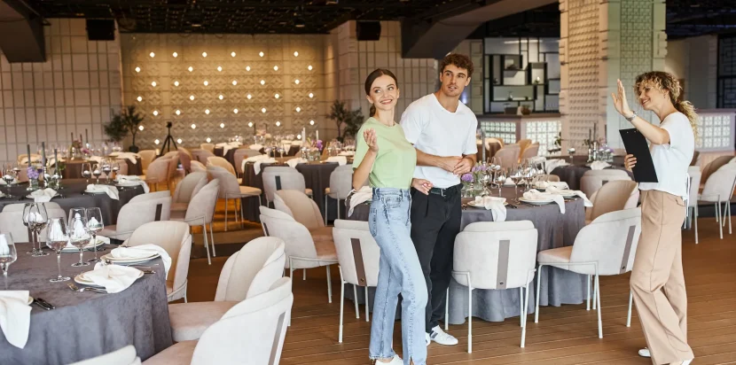 Grupo de personas en un restaurante elegante, con mesas bien decoradas y vajilla lista para una ocasión especial. Dos personas posan sonrientes, mientras una tercera les muestra un menú, creando un ambiente acogedor y moderno.