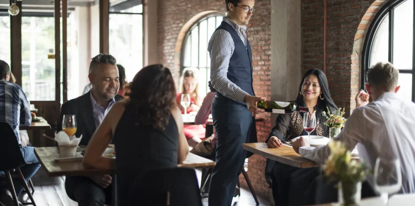 Cliente disfrutando de una cena en un restaurante moderno, mientras un camarero sirve vino a una mesa. Ambiente acogedor con decoración de ladrillo expuesto y luz natural.