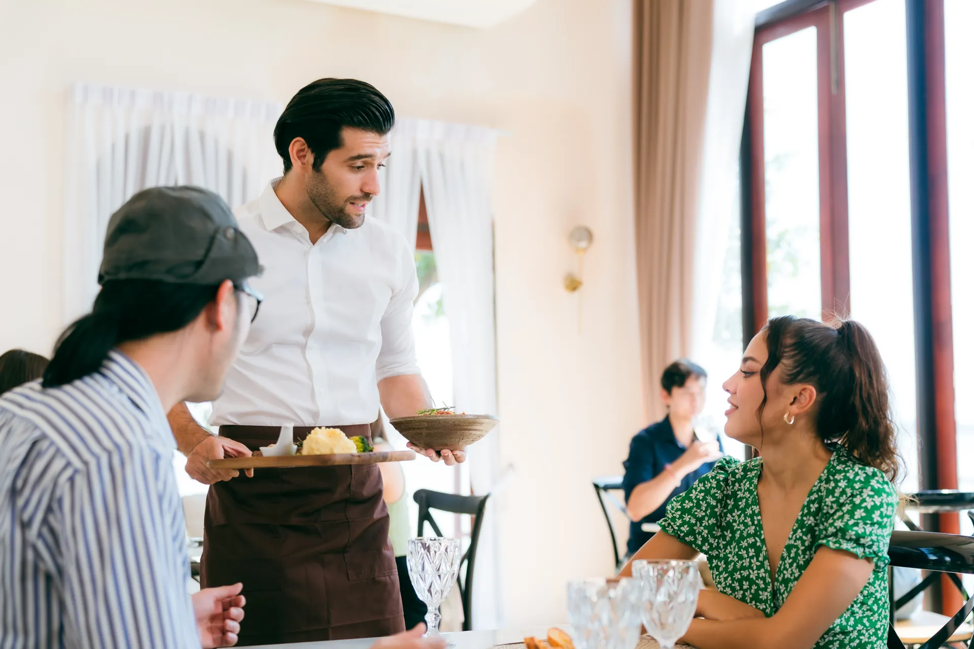 Camarero sirviendo un plato a una mujer en un restaurante, con otros comensales en el fondo. La decoración es moderna y luminosa, creando un ambiente acogedor.